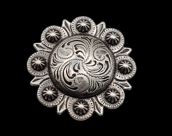 May include: A silver-toned metal concho with a floral design. The concho is circular in shape with a raised centre and a scalloped edge.