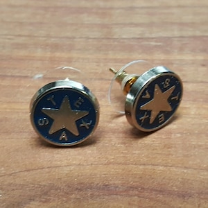 May include: A pair of gold-toned earrings with a blue enamel background and a gold star in the center. The star is surrounded by the letters "TEXAS" in blue enamel.