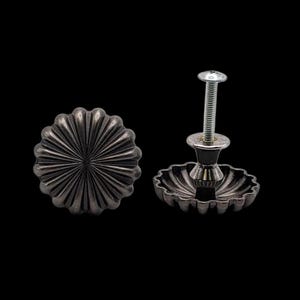 May include: Two decorative, dark gray metal cabinet knobs with a fluted, flower-like design. One knob is separate, and the other has a screw attached. The knobs are set against a black background.
