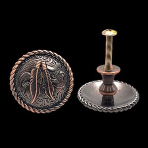 Pode incluir: Uma maçaneta de armário de cobre e preto com um design de corda ao redor da borda. A maçaneta apresenta um design em relevo de duas penas com um padrão floral.