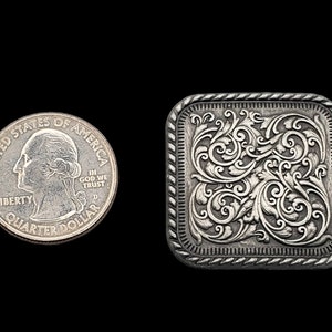 Op de afbeelding: Een zilverkleurige, vierkante gesp met een sierlijk bloemmotief. De gesp staat naast een Amerikaanse kwart dollar ter vergelijking van de grootte. De gesp heeft een touwachtige rand en staat tegen een zwarte achtergrond.
