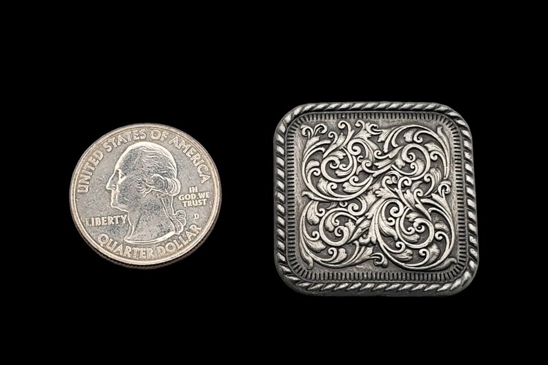 Op de afbeelding: Een zilverkleurige, vierkante gesp met een sierlijk bloemmotief. De gesp staat naast een Amerikaanse kwart dollar ter vergelijking van de grootte. De gesp heeft een touwachtige rand en staat tegen een zwarte achtergrond.