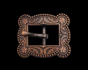 May include: A rectangular copper-toned belt buckle featuring an ornate border with embossed floral motifs and a beaded edge. The center contains a rectangular opening with a horizontal prong, showcasing a dark, weathered finish in the recessed areas.