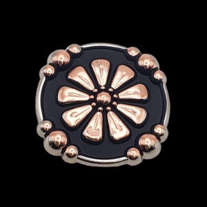 May include: A round, decorative button with a black center and a floral design in rose gold. The button is surrounded by a silver border and small, rose gold spheres.