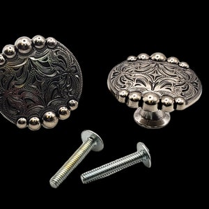 May include: Two silver-toned, round, decorative cabinet knobs with a floral design and a row of raised beads around the edge. The knobs are shown with two silver screws.