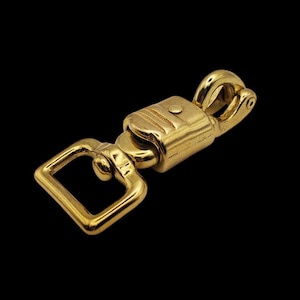 Op de afbeelding: Een gouden metalen trekker-snelkoppeling met een vierkante lus aan het ene uiteinde en een draaibare haak aan het andere uiteinde.