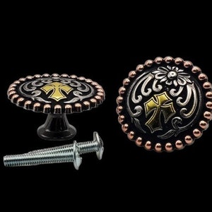 May include: Decorative cabinet knobs with a Western-inspired design. The knobs feature a black base with silver and gold accents, a cross design, and a copper-coloured beaded edge. Includes a screw for installation.