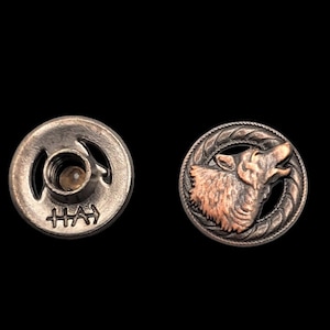 May include: A pair of metal jean buttons. One button is silver with a circular design and the text "HHA" on the bottom. The other button is copper with a wolf's head design.