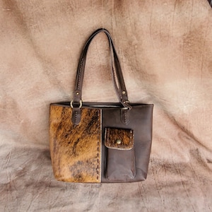 May include: A brown leather tote bag with a brown and tan cowhide patch on the front. The bag has a zip closure and a small pocket on the front.