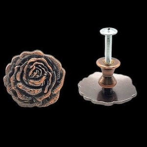 May include: A copper-toned rose-shaped cabinet knob with a silver screw. The knob is attached to a round base with a scalloped edge.