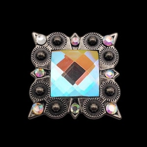 May include: A square-shaped, silver-toned metal buckle with a large, iridescent, faceted crystal in the center. The crystal has a blue, green, and orange color scheme. The buckle is decorated with smaller, clear crystals around the edge.