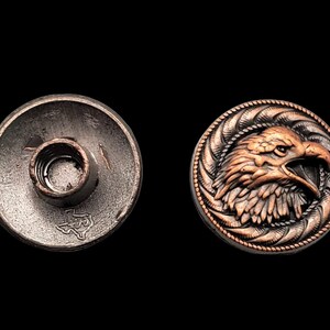 May include: A copper-toned metal button with an eagle head design. The button has a detailed, textured design and a raised eagle head with a sharp beak. The back of the button is plain metal.