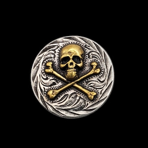 Op de afbeelding: Een zilveren en gouden knoop met een schedel en gekruiste beenderen met een gedetailleerd gegraveerd ontwerp. De schedel is goud en de gekruiste beenderen zijn goud op een zilveren achtergrond.