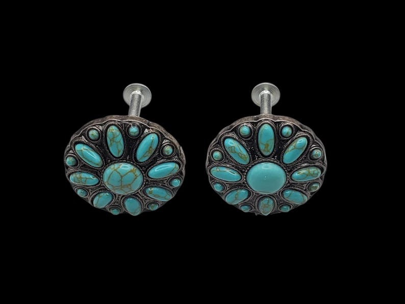 Może przedstawiać: Dwa dekoracyjne gałki do szafek ze srebrnym wykończeniem i turkusowymi kamiennymi akcentami. Gałki mają kwiatowy wz&oacute;r z dużym centralnym kamieniem i mniejszymi kamieniami na obwodzie. Zamontowane na srebrnych śrubach.