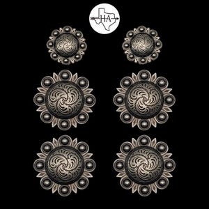May include: Six circular silver-toned metal conchos arranged in three pairs of varying sizes. Each piece features a central engraved swirl pattern surrounded by a border of small raised spheres and pointed petals. A circular logo with the text HA is centered above.