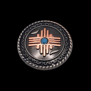 Puede incluir: Un botón de metal de color plateado y cobre con una piedra turquesa en el centro y un símbolo de sol estilizado con ocho rayos.