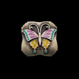 Pode incluir: Broche de metal prateado em forma de borboleta. A borboleta tem asas rosa e amarelas com detalhes turquesa. A broche está sobre um fundo escuro.