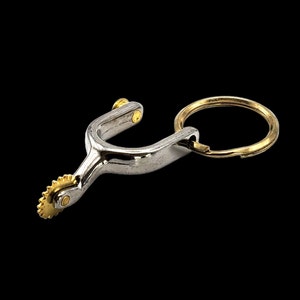 May include: A silver metal spur keychain with a gold-colored ring and a decorative wheel. The spur is a traditional cowboy accessory.