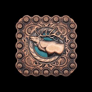 May include: A copper-colored belt buckle with an ornate design featuring an elk with antlers in a circular frame. The elk is in relief and has a turquoise background.