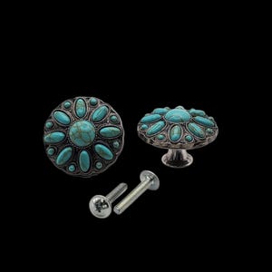 Może przedstawiać: Dekoracyjne gałki do szafek ze srebrnym wykończeniem i turkusowymi kamieniami. Gałki mają kwiatowy wz&oacute;r i są dostarczane ze śrubami montażowymi. Gałki są pokazane na czarnym tle.