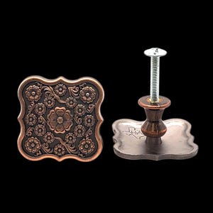 May include: Two decorative cabinet knobs with a floral design. The knobs are square with a scalloped edge and a copper finish. One knob is shown with a screw and a dark brown wooden base.