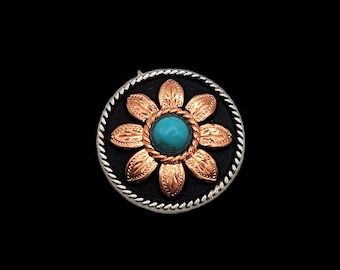 May include: A silver and copper colored flower-shaped button with a turquoise stone in the center. The button has a black background and a silver rope-like border.