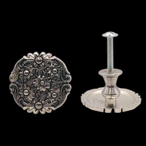 May include: Two silver-toned cabinet knobs with floral designs. One knob is a decorative, round plate with a detailed flower pattern. The other knob has a screw and a decorative base. The knobs are set against a black background.