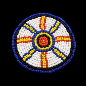 May include: A round beaded design with a white background and concentric circles of blue, black, yellow, and red beads.