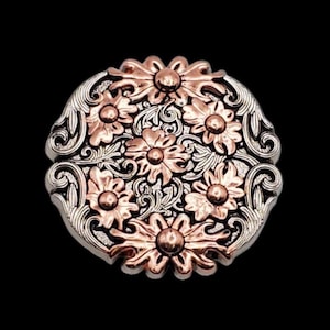 Puede incluir: Hebilla metálica redonda con diseño floral en tonos plateados y cobrizos. Las flores están dispuestas simétricamente, con un borde grabado detallado. La hebilla se encuentra sobre un fondo negro.