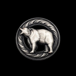 Puede incluir: Un botón de metal plateado con un diseño detallado de un oso en un círculo. El oso está mirando hacia la derecha y tiene un diseño texturizado detallado.