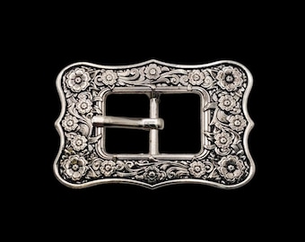 May include: Silver-toned metal belt buckle with an ornate floral design. The rectangular buckle features a central bar and a decorative frame with detailed flower and leaf patterns. The background is solid black.