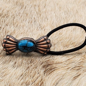 May include: A turquoise and copper hair tie with a decorative bow design. The bow features a turquoise stone in the center.