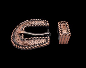 May include: A copper-toned belt buckle with an intricate floral design and a black metal buckle. The buckle is accompanied by a matching copper-toned belt tip.