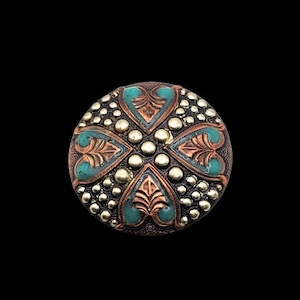 Puede incluir: Botón redondo de estilo antiguo con un diseño detallado. El botón presenta una base de color cobre con detalles turquesa y dorados. El diseño incluye un patrón en forma de cruz con detalles de hojas y puntos.