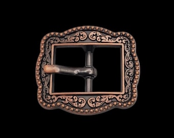 May include: A rectangular belt buckle featuring a dark metallic finish with copper-toned highlights. The frame is decorated with a beaded outer edge and an intricate scrollwork pattern. A central prong extends horizontally across the open center.