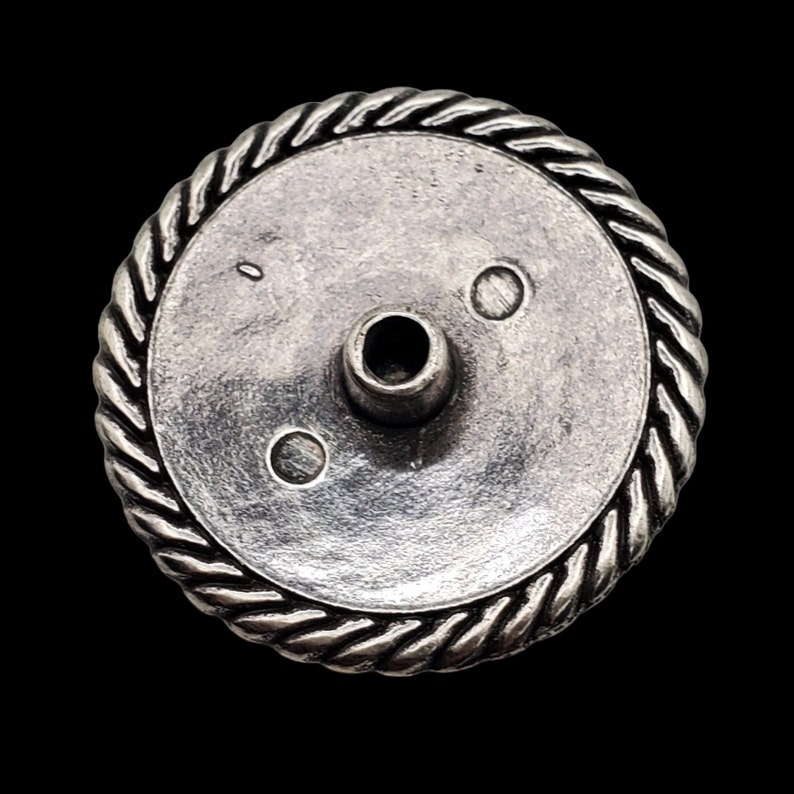 May include: Silver-toned metal button with a rope-like design around the edge. The button has a small hole in the center and three small circles on the surface.