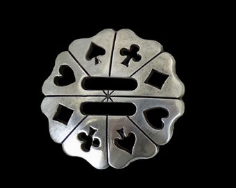 May include: A silver-toned metal pendant with a circular design featuring cutouts of playing card suits. The pendant has a scalloped edge and is decorated with a cross-shaped design in the center.