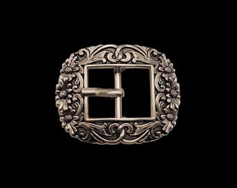 May include: A rectangular silver-toned metal belt buckle featuring an ornate border with embossed floral motifs and scrolling patterns. The center contains a square opening with a horizontal prong mechanism for securing a belt strap.