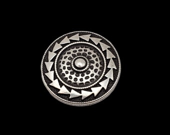 Op de afbeelding: Een zilverkleurige metalen knoop met een cirkelvormig ontwerp. De knoop heeft een centrale cirkel die wordt omringd door een ring van kleinere cirkels, allemaal binnen een grotere cirkel met pijlvormige vormen.