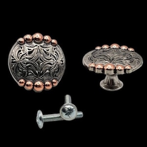 Pode incluir: Um conjunto de duas maçanetas de armário de prata e cobre com um design floral. As maçanetas têm uma base redonda e uma parte superior elevada e adornada. As maçanetas são presas a um parafuso de prata.