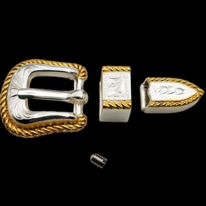 Könnte beinhalten: Eine silberne Gürtelschnalle mit einem goldenen Seil-Design am Rand. Die Schnalle ist mit einem floralen Muster graviert. Die Schnalle wird mit einer passenden Spitze und einer Schraube geliefert.