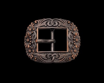 May include: A rectangular belt buckle with a dark copper finish, featuring an ornate relief pattern of swirling scrollwork and floral motifs along the border. The center contains a standard rectangular frame with a central prong mechanism.