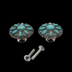 Może przedstawiać: Dwa dekoracyjne gałki do szafek ze srebrnym wykończeniem i inkrustowanymi turkusowymi kamieniami. Gałki mają kwiatowy wz&oacute;r i towarzyszą im dwie srebrne śruby, wszystko na czarnym tle.