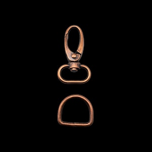 Könnte beinhalten: Ein kupferfarbener Metall-Drehverschluss und ein D-Ring. Der Verschluss hat einen federbelasteten Verschluss und eine Schlaufe am anderen Ende. Der D-Ring ist eine einfache Schlaufe.
