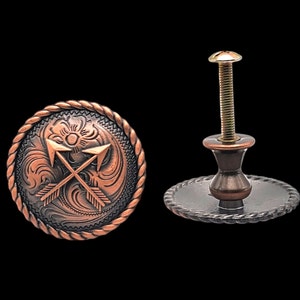 Pode incluir: Uma maçaneta de armário em tom de cobre com um design decorativo de duas flechas cruzadas e um padrão floral. A maçaneta tem uma base redonda e um parafuso roscado.