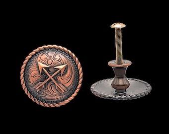 Puede incluir: Un pomo de armario de tono cobre con un diseño decorativo de dos flechas cruzadas y un patrón floral. El pomo tiene una base redonda y un tornillo roscado.