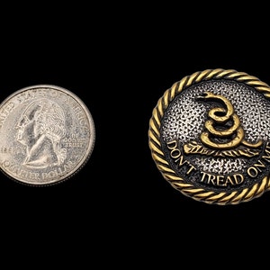 May include: A gold-colored metal button with a black and white textured background. The button features a gold-colored snake with the text "Don't Tread On Me" around the snake.