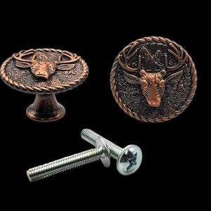 May include: Two copper-toned cabinet knobs featuring a deer head design. The knobs are round and have a rope-like border. One knob is attached to a base, the other is not. A silver screw is also shown.