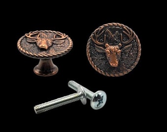 Puede incluir: Dos pomos de armario de color cobre con un diseño de cabeza de ciervo. Los pomos son redondos y tienen un borde de cuerda. Un pomo está unido a una base, el otro no. También se muestra un tornillo plateado.