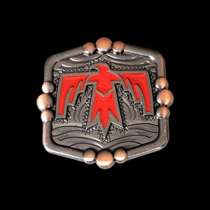 May include: A silver-toned belt buckle with a red stylized bird design. The buckle has a geometric pattern around the bird and a textured surface.